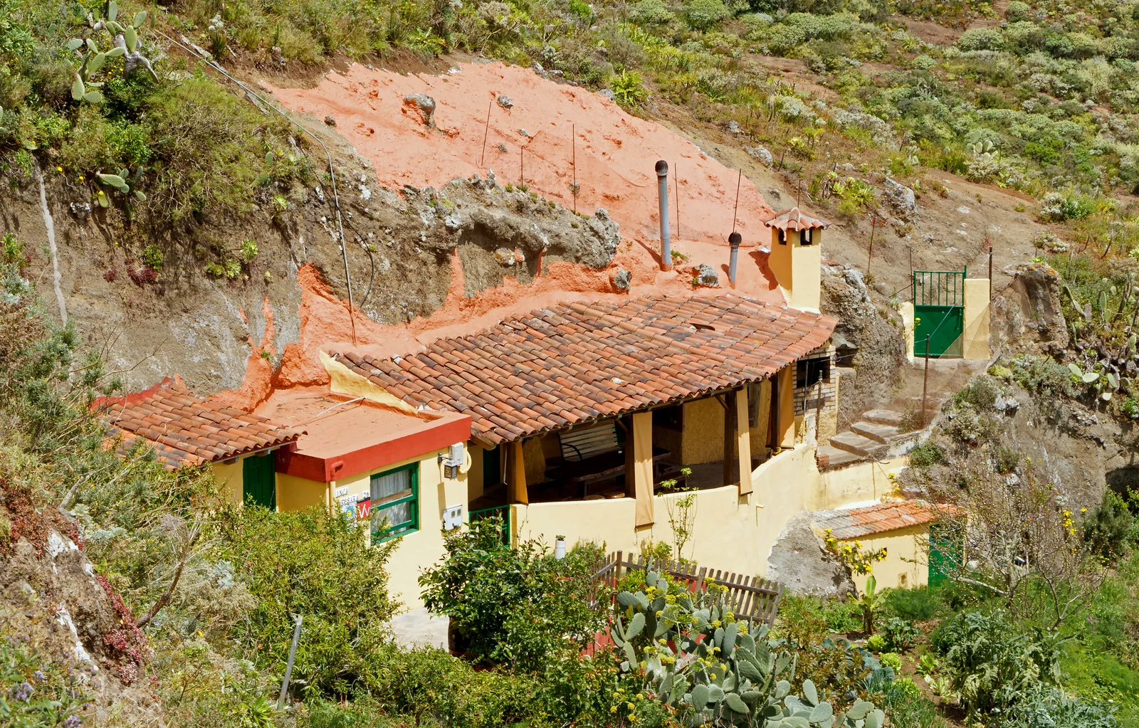 Casa cueva del caserío de Chinamada, en el macizo de Anaga (Tenerife): vivienda de fachada amarilla con zócalo rojo y tejado de teja árabe adosada a la ladera, con la cavidad excavada en la toba volcánica a la derecha, chimeneas asomando por el terreno sobre la cubierta y bancales con tuneras alrededor