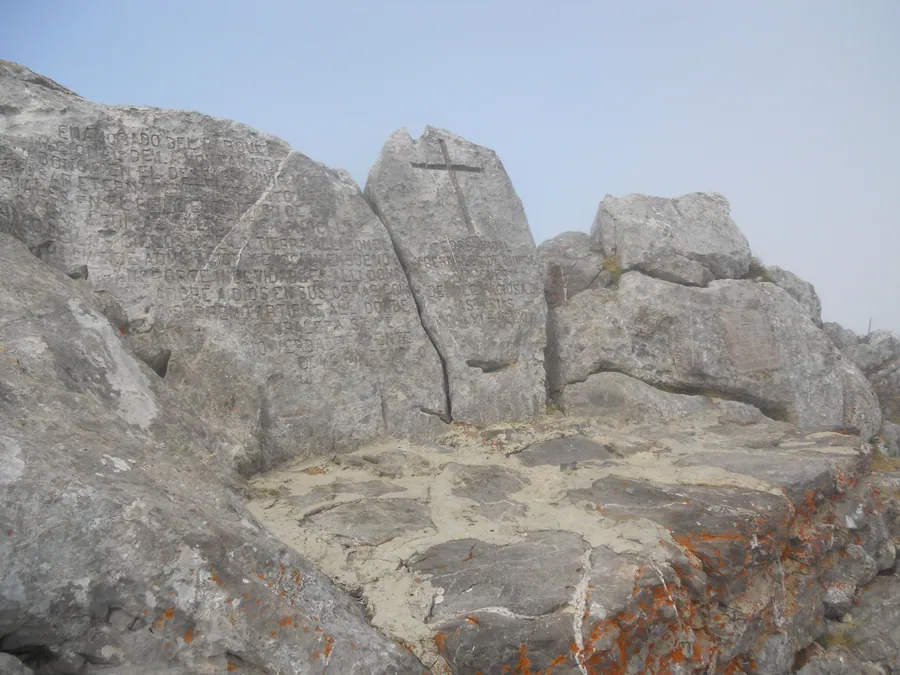 La tumba de Pedro Pidal en el mirador de Ordiales, con el epitafio grabado en la roca caliza y una cruz tallada en el bloque contiguo, bajo un cielo claro con bruma, en el macizo del Cornión, Parque Nacional de los Picos de Europa, Cangas de Onís (Asturias)